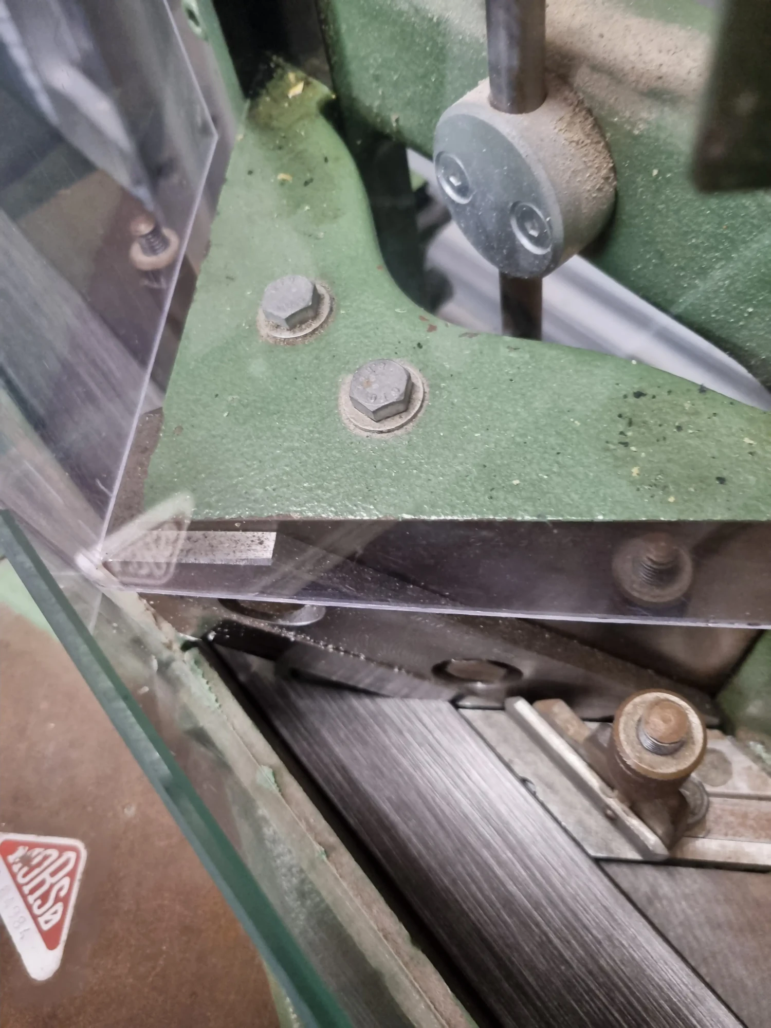 Close-up of an industrial machine with green metal parts, bolts, and screws, partially covered by a clear protective shield. Ideal for crafting Handmade Bespoke Picture Frames, the machine features moving parts and cutting or pressing components.