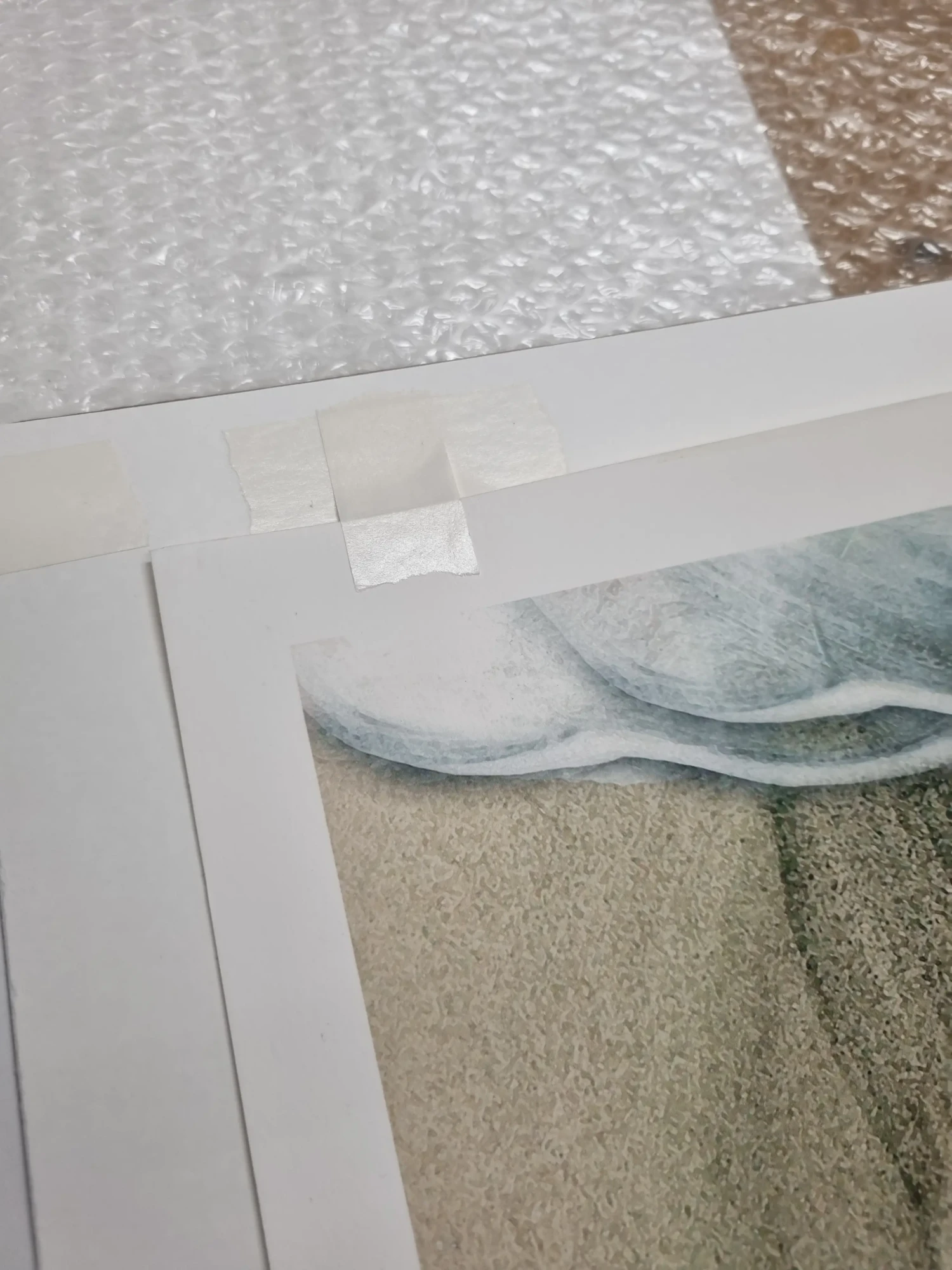 A close-up of two pieces of white paper taped together at the top edge with masking tape; one paper has part of a drawing showing light blue and beige shades. Bubble wrap and handmade bespoke picture frames are visible in the background.
