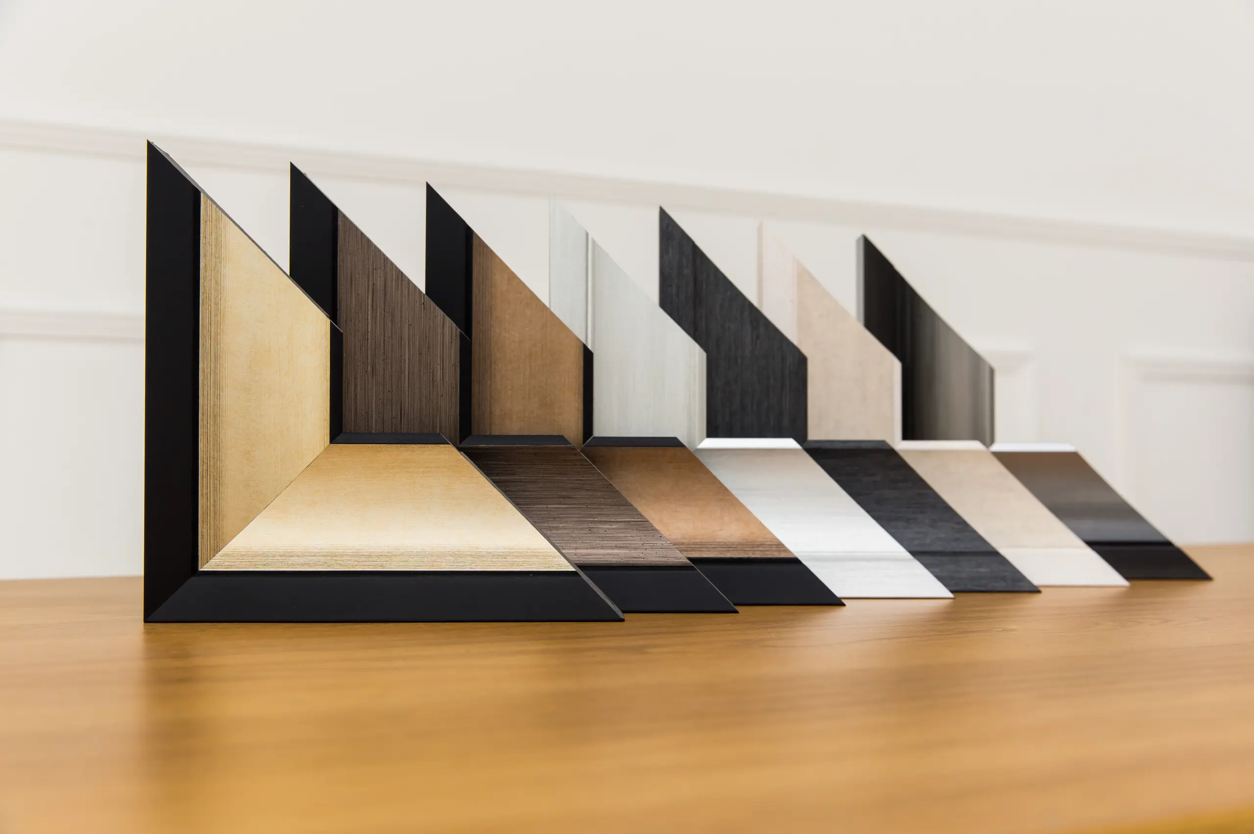 A row of polymer picture frame corners in various colours and finishes is displayed on a light wooden surface, with each frame angled upright against a white background.