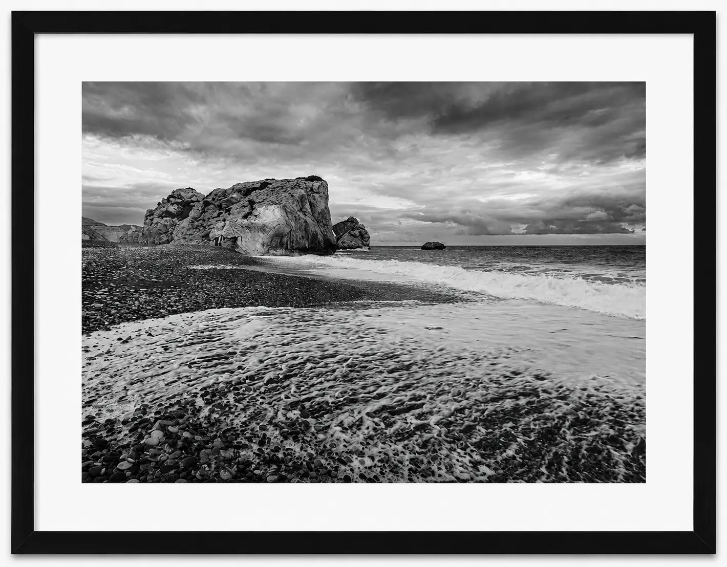 Black and white photo of a rocky beach with waves washing ashore, a large rock formation in the background, dramatic clouds overhead, and a framed border—part of the striking Noel Fenech Framed Prints collection.
