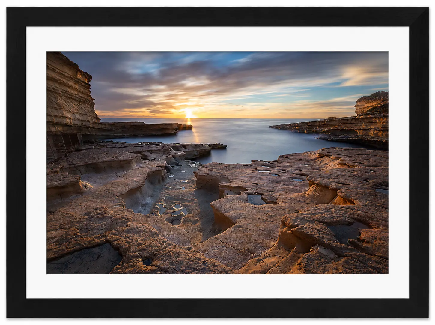 A coastal landscape at sunset, with layered rock formations in the foreground and the sun shining over a calm sea. Dramatic clouds fill the sky in this striking black and white image, available as Noel Fenech Framed Prints.