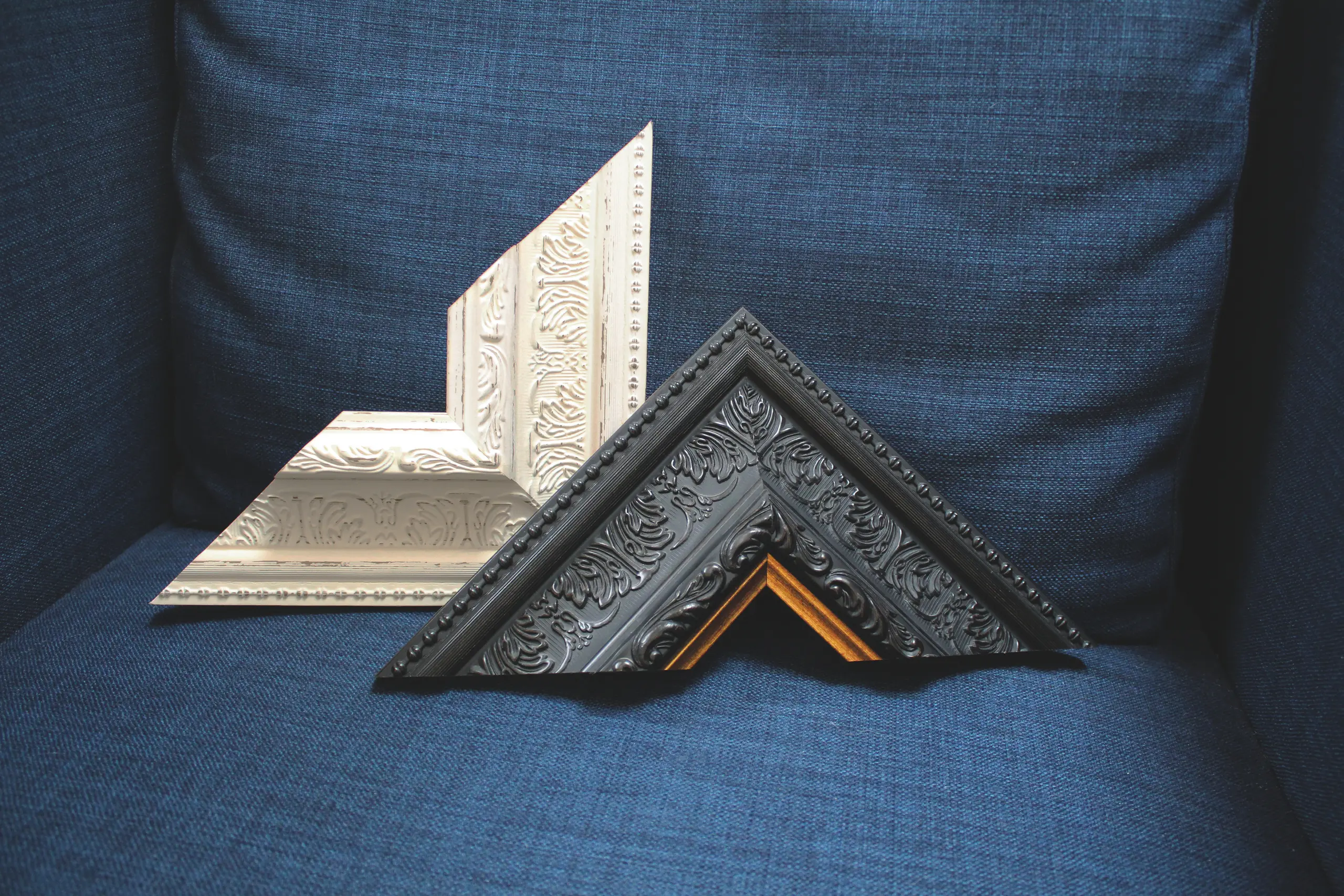 Two ornate polymer picture frame corners, one white and one black with gold, rest on a dark blue fabric chair.