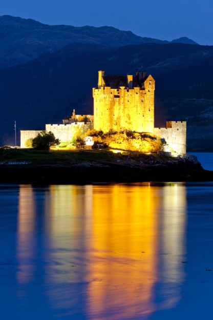 Eilean Donan Castle illuminated at night, reflecting on Loch Duich in a portrait-oriented photograph