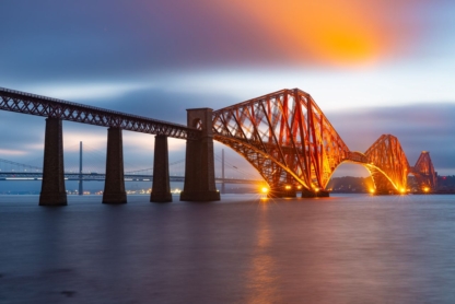 A tranquil evening view of the Forth Bridge, a railway bridge spanning the Firth of Forth near Queensferry, its illuminated red lattice structure reflected in the calm waters.